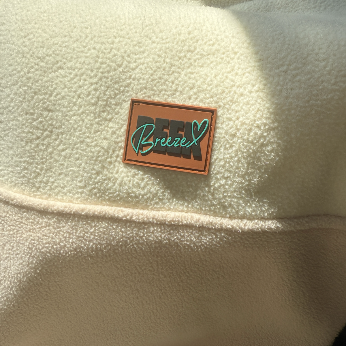 Close-up of the signature Peek Breeze brown logo patch on the soft ivory fleece fabric of the Oasis women’s quarter-zip dress.