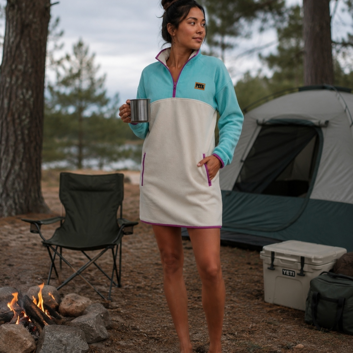 Woman holding a mug by a campfire wearing the Breeze fleece dress in turquoise and beige with pink trim